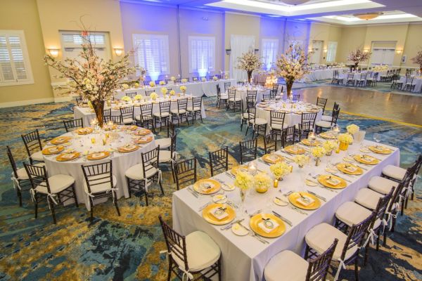 The image shows a large, elegantly decorated banquet hall with dining tables set for an event, adorned with floral centerpieces and white tablecloths.