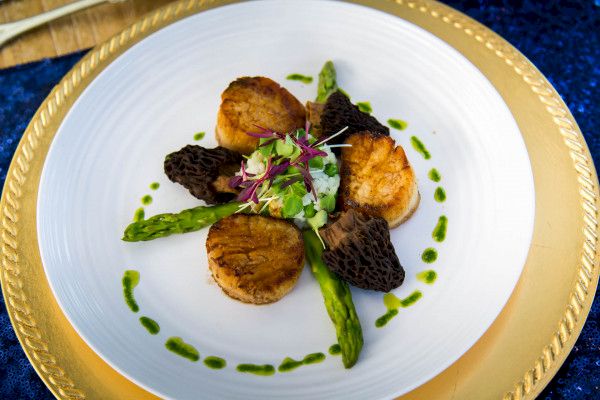 The image shows a gourmet dish with seared scallops, asparagus spears, morel mushrooms, and a garnish of microgreens, plated on a gold-rimmed dish.