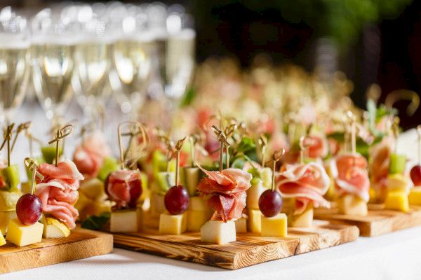 The image shows a display of appetizers on wooden boards, featuring cheese, grapes, and cured meat, with glasses of champagne in the background.