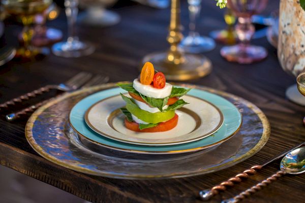 A beautifully plated salad with tomatoes, cheese, and greens is arranged in a stack on a decorative plate setting on a wooden table.