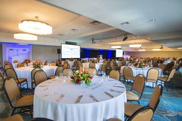 A large conference room with round tables set for dining, floral centerpieces, presentation screens, and rows of chairs, likely set for an event.