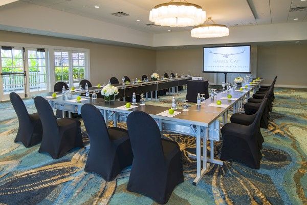 A conference room with a U-shaped table setup, black chair covers, water bottles, notepads, centerpieces, a projector screen, and large windows.