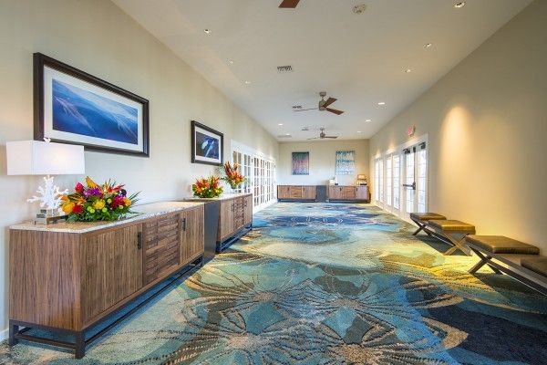 A brightly lit hallway with blue patterned carpet, wooden sideboard, flowers, lamps, framed pictures, ceiling fans, and benches.