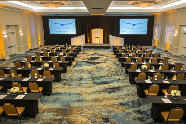 A conference room set up with rows of tables and chairs, a stage with a podium, and two large screens displaying "Hawk’s Cay".