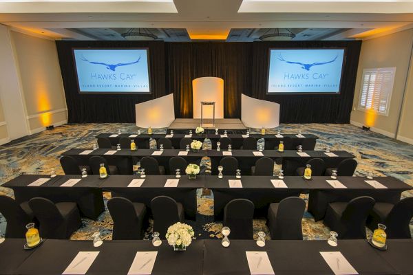 This image shows a conference or meeting room setup with rows of tables and chairs, two projection screens, and a podium in the center.