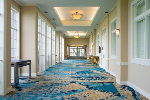 A well-lit hallway with large windows on one side and closed doors on the other, featuring blue-patterned carpeting and overhead chandeliers.