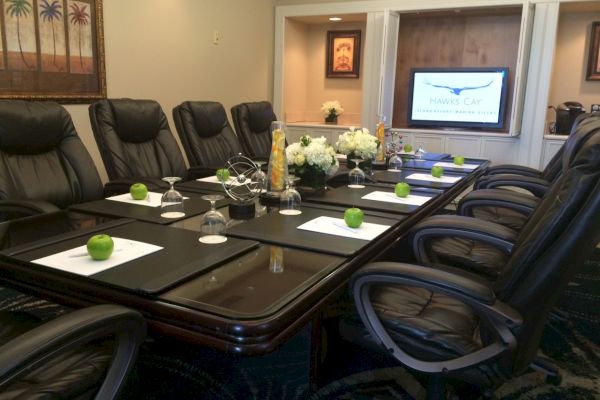 A conference room with a long table, black chairs, apples, wine glasses, notepads, and a screen displaying "Meeting Room." Ending the sentence.