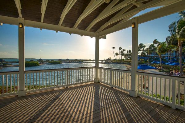 A scenic view from a gazebo overlooking a serene beach and palm trees at sunset, with a wooden deck and shadows from the railing and roof structure.