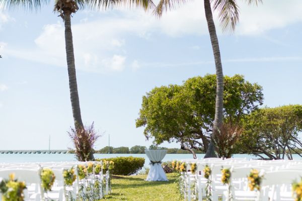 An outdoor wedding setup with rows of white chairs decorated with yellow flowers, situated between two palm trees by the water under a blue sky.