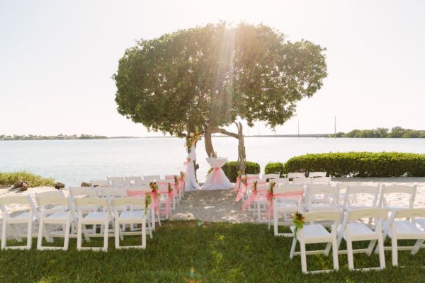 An outdoor wedding setup with rows of white chairs lined up facing a decorated arch and table under a tree by a water body, on a sunny day.