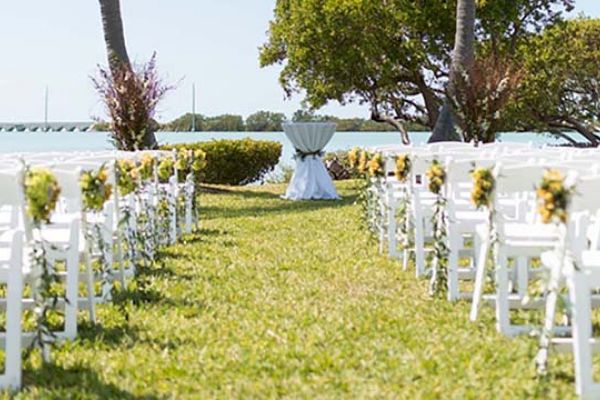 An outdoor wedding setup has white chairs adorned with flowers, forming an aisle leading to a small table, with the ocean and trees in the background.