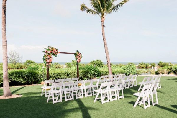 An outdoor wedding setup with white chairs, a floral arch, and a backdrop of greenery and the ocean, under palm trees.