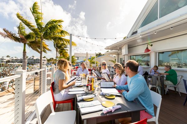 A group of people are dining at an outdoor restaurant by the waterfront, surrounded by palm trees and string lights, enjoying a sunny day.