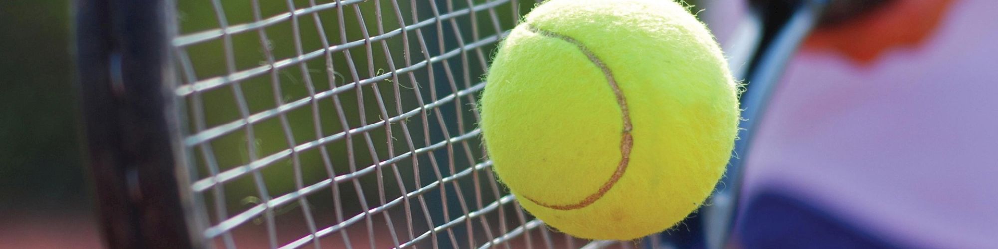 A close-up image of a tennis ball in motion about to hit the strings of a tennis racket on a tennis court.