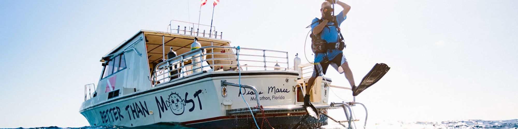 A person is jumping into the water from a boat with diving gear. The boat has writing on the side that reads "BETTER THAN MOST."