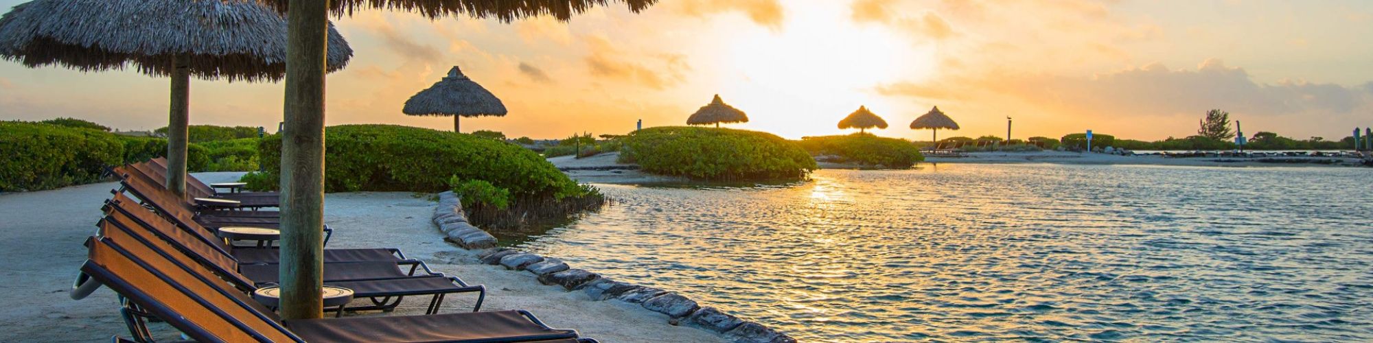 A serene beach setting with lounge chairs under thatched umbrellas facing a tranquil body of water during a picturesque sunset.