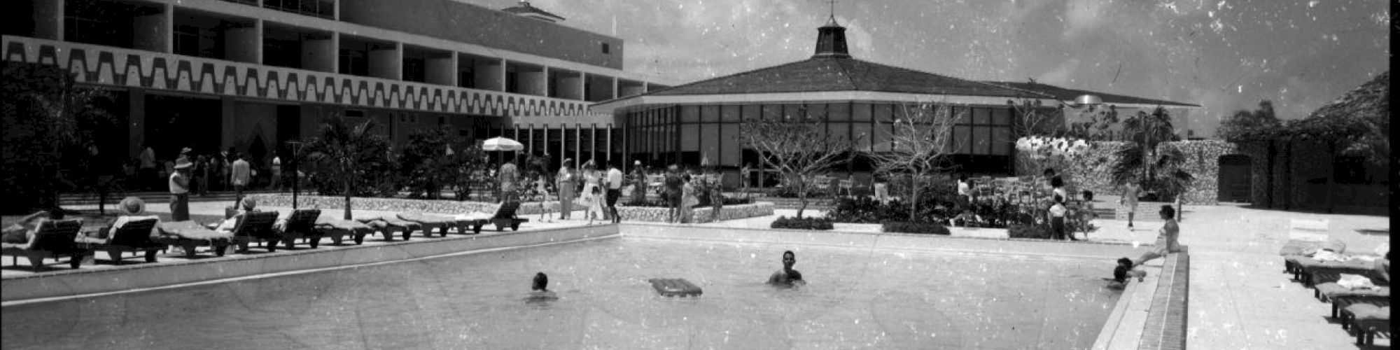 The image shows a vintage scene of a hotel pool area with several people swimming and sunbathing, with the hotel building in the background.