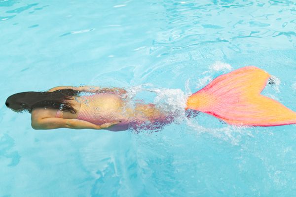 A person wearing a mermaid costume with a pink and orange tail is swimming underwater in a pool, creating a whimsical and fantasy-like scene.