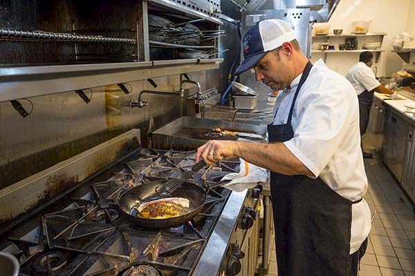 A chef in a commercial kitchen is cooking food in a pan on a stovetop, with another chef working in the background.