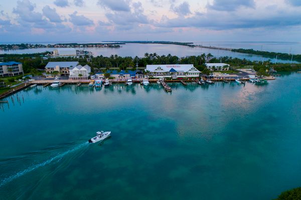 An aerial view of a coastal town with buildings near the water, a boat sailing, docks, light clouds, and distant bridges on a tranquil day.