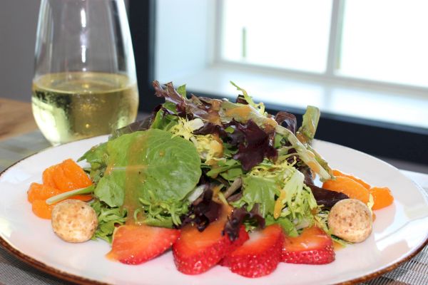 A fresh salad with mixed greens, strawberries, mandarin slices, and cheese balls sits next to a glass of white wine on a table.