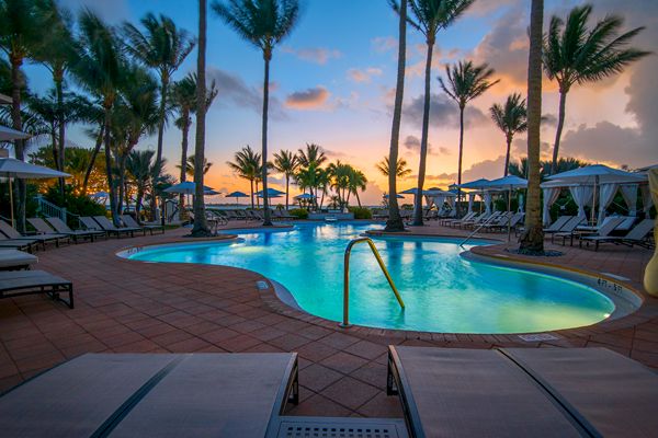 A luxurious pool area surrounded by lounge chairs and palm trees at dusk, with a colorful sky in the background, creating a serene setting.