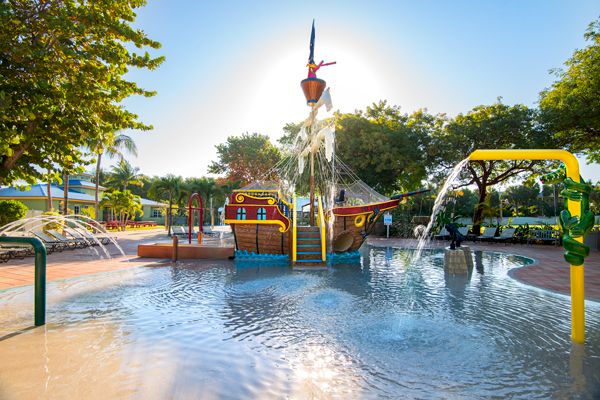 The image shows a children's splash pad with a pirate ship-themed water play structure, surrounded by trees, water sprayers, and clear blue skies.