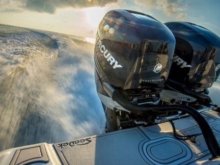 The image shows two Mercury outboard motors on a boat moving swiftly across the water, creating a wake behind them as the sun sets in the background.