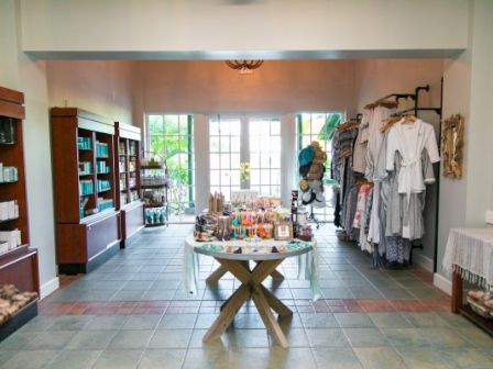 A boutique store with shelves of products, clothing racks, and a central display table, all arranged in a well-lit room with tiled flooring.