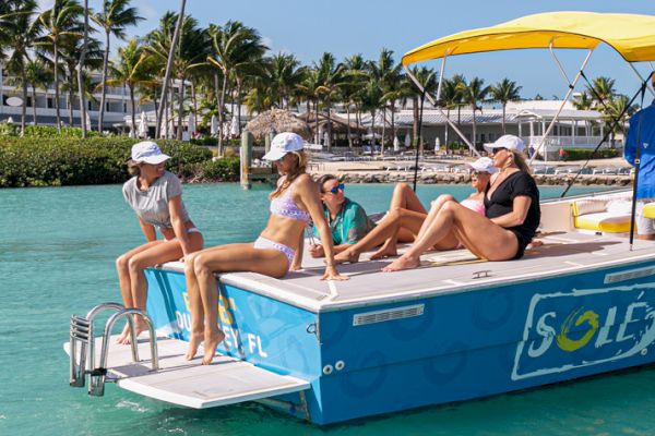 A group of people on a blue "Sole Watersports" boat with a yellow canopy, enjoying the water near a tropical resort.