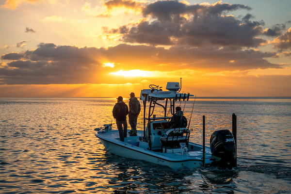 A boat with three people on a calm body of water at sunset, with the sun casting rays through clouds in the sky.