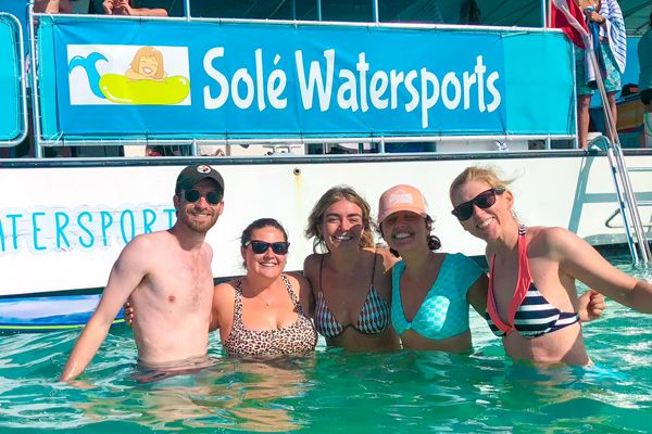 Five people smiling in a body of water in front of a sign that reads "Solé Watersports" attached to a boat, enjoying a sunny day.