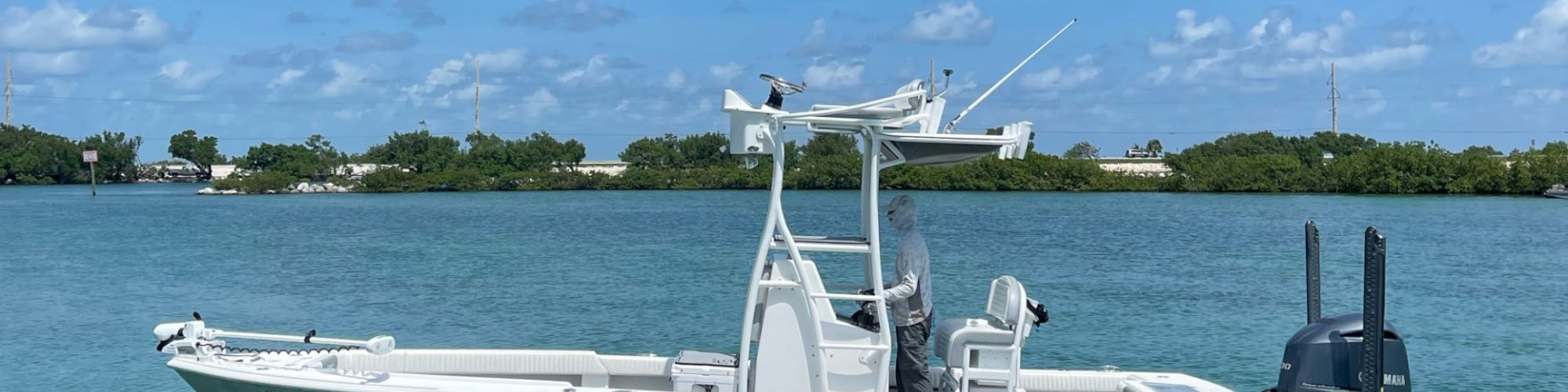 A white fishing boat with an outboard motor is floating on calm water with green foliage and a blue sky visible in the background.