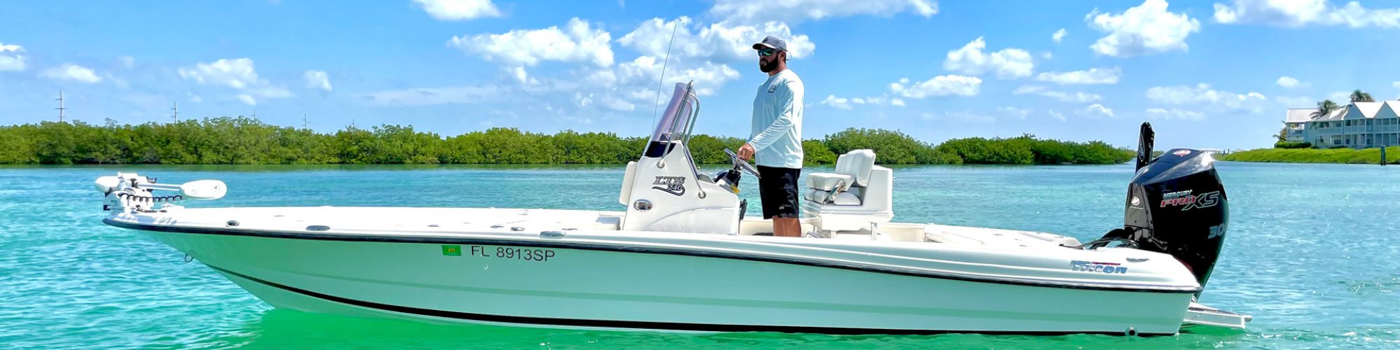 A person stands on a small white motorboat on clear turquoise water under a bright blue sky with scattered clouds.