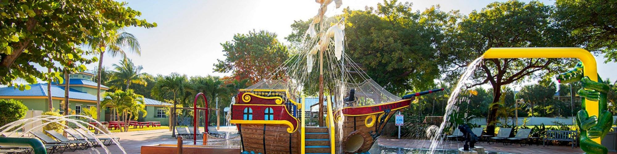 A children's water play area featuring a pirate ship with water slides and fountains, surrounded by trees and seating areas, in sunlight.