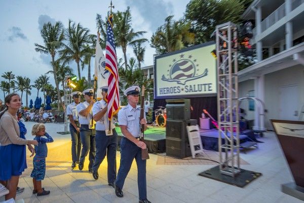 People in uniform are carrying flags in a procession next to a "Heroes Salute" sign, with onlookers nearby and palm trees in the background.