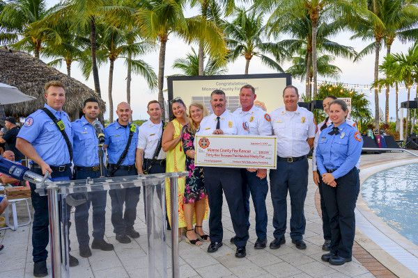 A group of people in uniform are standing around a large check at an outdoor event with palm trees in the background, next to a pool.