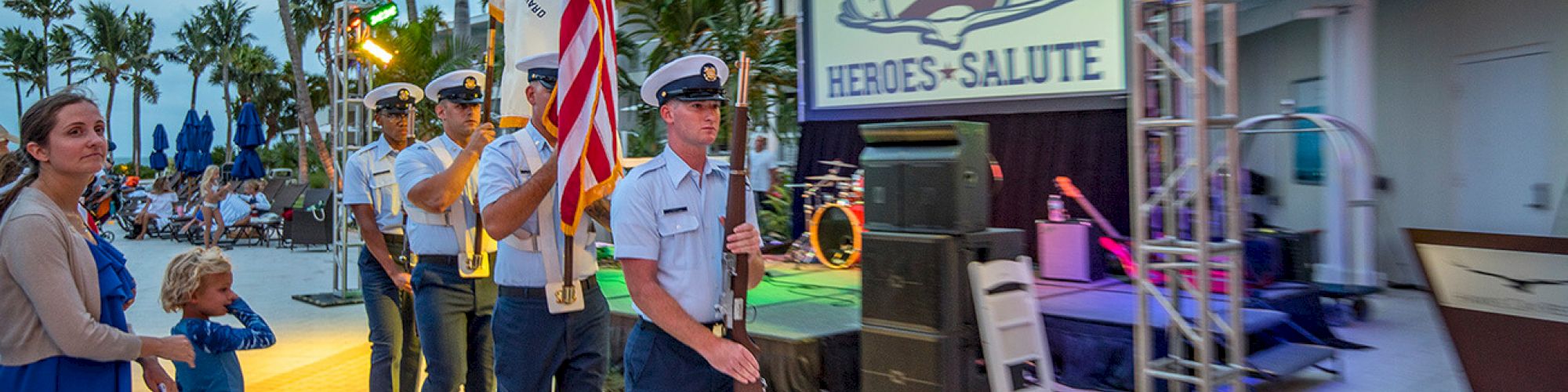 A group of uniformed individuals bearing flags march at an outdoor event titled "Heroes Salute"; a woman and child watch from the side.