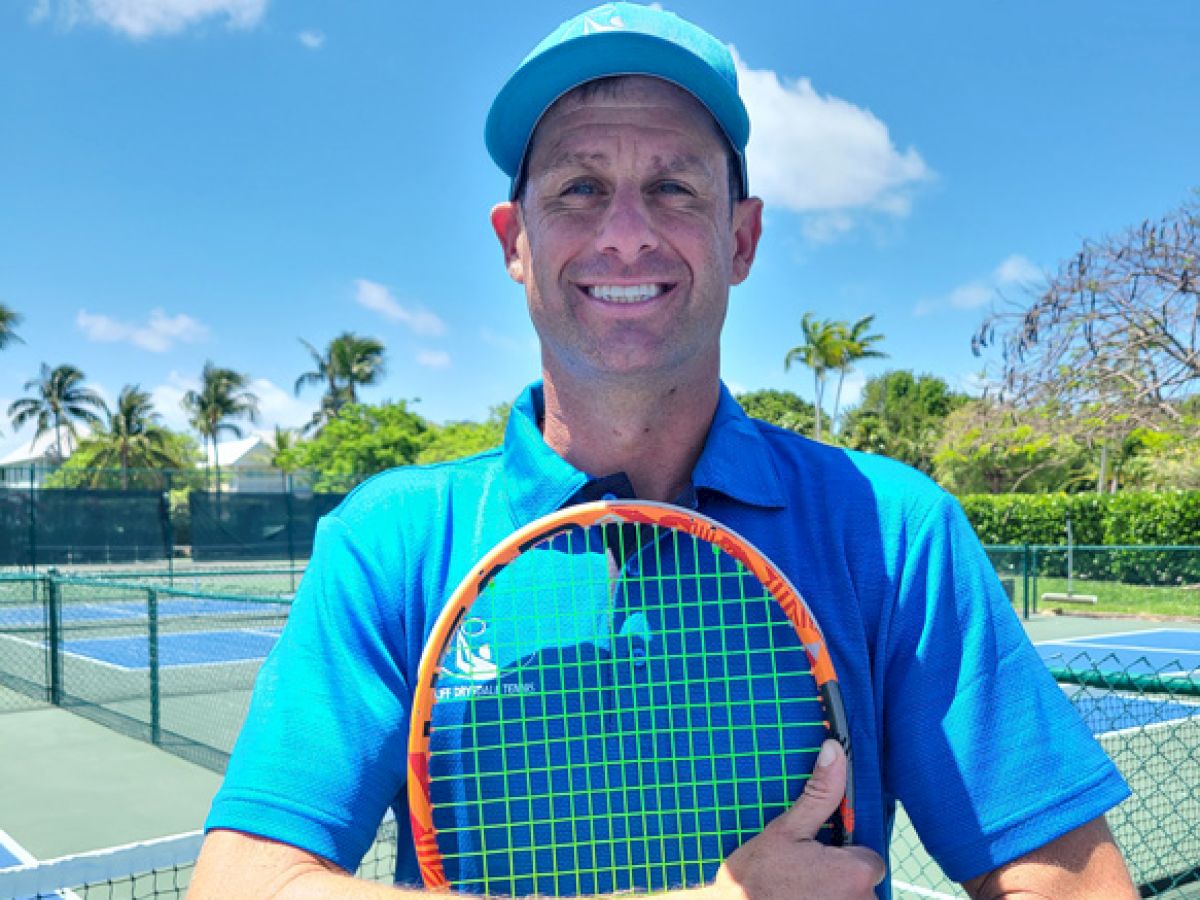 A smiling man in a blue shirt and cap holds a tennis racket on an outdoor court with palm trees and a clear sky in the background.