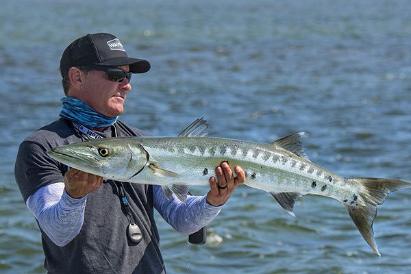 A person, wearing a hat and sunglasses, is holding a large fish near a body of water, possibly indicating a fishing activity or catch.