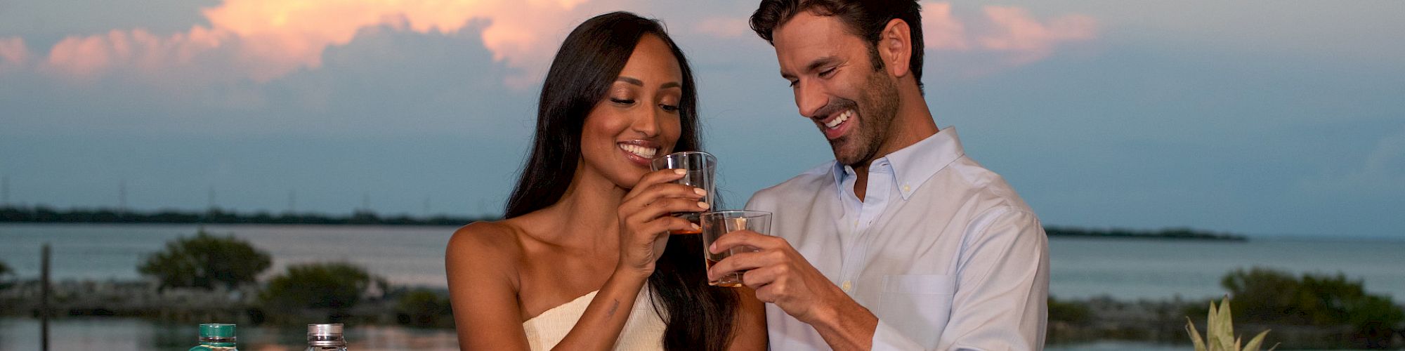 A couple toasts with drinks near a waterfront. Multiple bottles, fruit-filled glasses, and a white bucket with "Key" text are on the table.