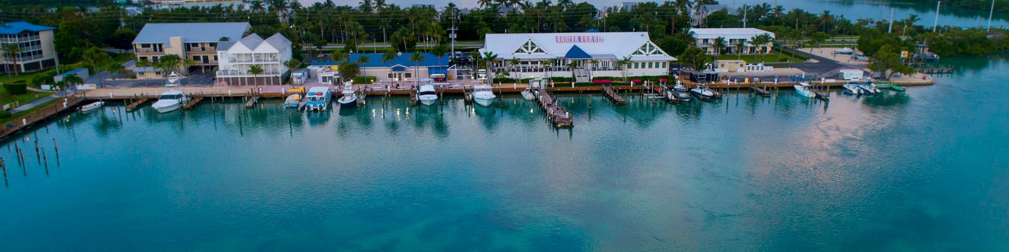 A coastal marina with boats docked, surrounded by buildings and lush trees, while a small boat navigates the calm blue water.