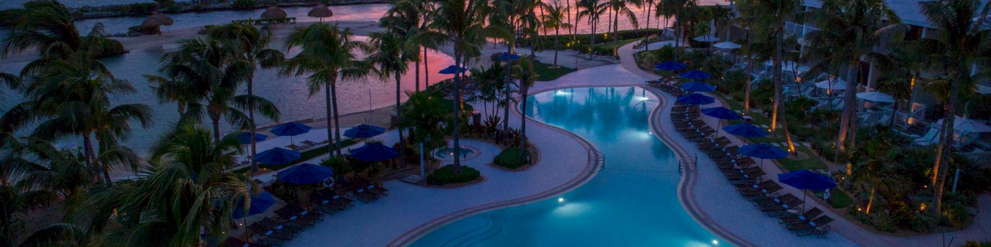 A resort pool at sunset with palm trees surrounding it, lounge chairs, and a beautiful ocean view in the background, under a colorful sky.