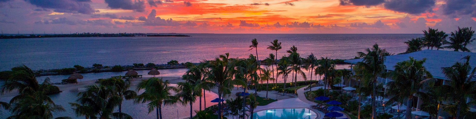 The image shows a luxurious resort with a large, winding swimming pool, surrounded by palm trees, with a vibrant sunset visible over the ocean.