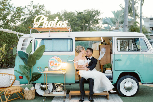 A bride and groom sit in a photo booth van with a retro design, surrounded by greenery and wedding decor, sharing an intimate moment together.