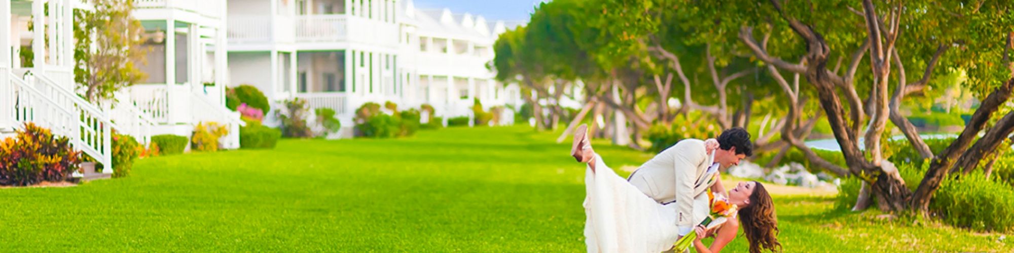 A couple in wedding attire dances joyfully on a lush lawn in front of white buildings and green trees, creating a serene and romantic scene.