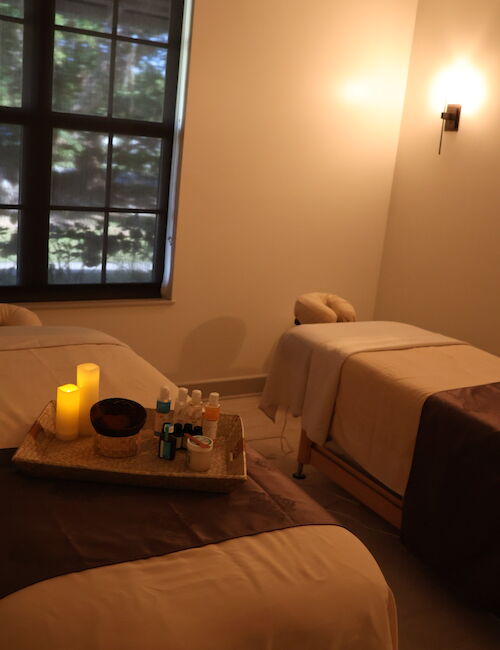 The image shows a cozy spa room with two massage tables, soft lighting, and lit candles on a tray, creating a relaxing atmosphere.