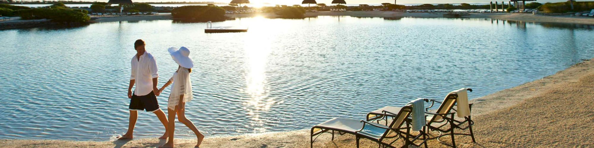 A couple is walking hand-in-hand along a beach near a calm lagoon, with lounge chairs and huts in the background under a bright, setting sun.