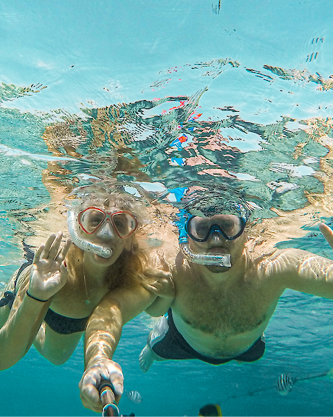 Two people snorkeling underwater, wearing masks and snorkels, taking a selfie. They seem to be in clear blue water.