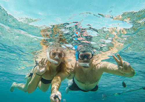 Two people snorkeling underwater, wearing masks and snorkels, taking a selfie. They seem to be in clear blue water.